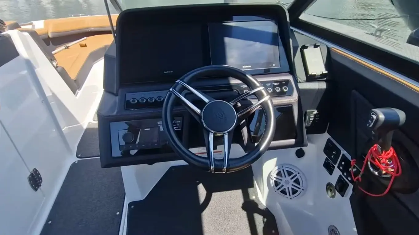 Boat cockpit featuring a steering wheel, dual screens, control panel, and audio system; light reflections on water visible.